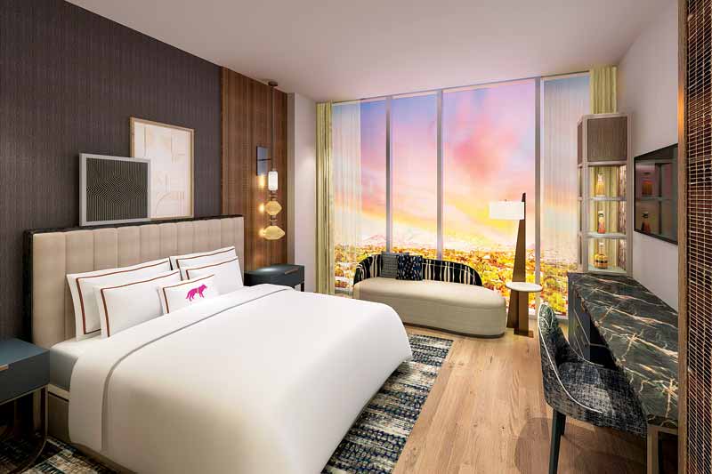 A guest room with a large bed positioned facing a desk, with an Ottoman placed against the window overlooking a view.