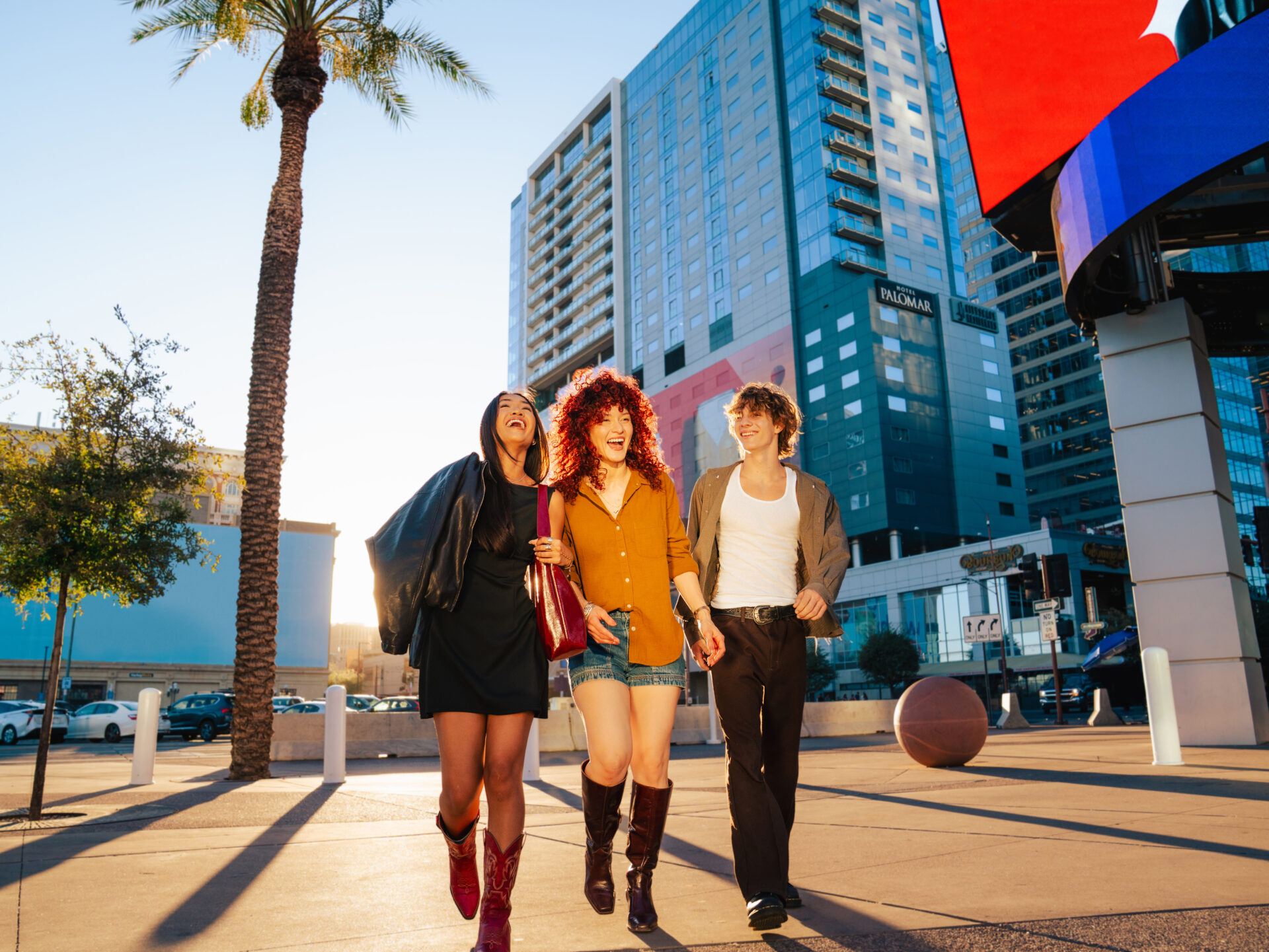 Three people walking arm in arm outside. There is a palm tree and sunlight in the background.