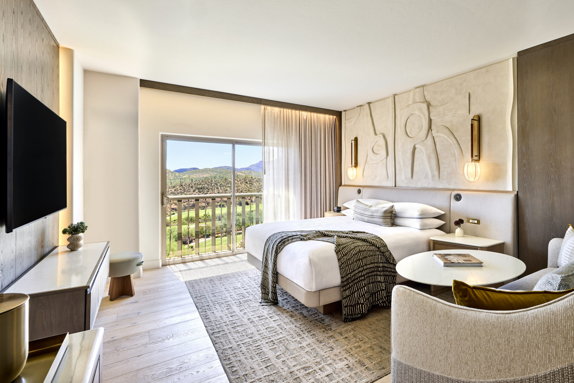 Guest room with a king bed, seating area, wall-mounted TV, and balcony overlooking a mountain landscape.