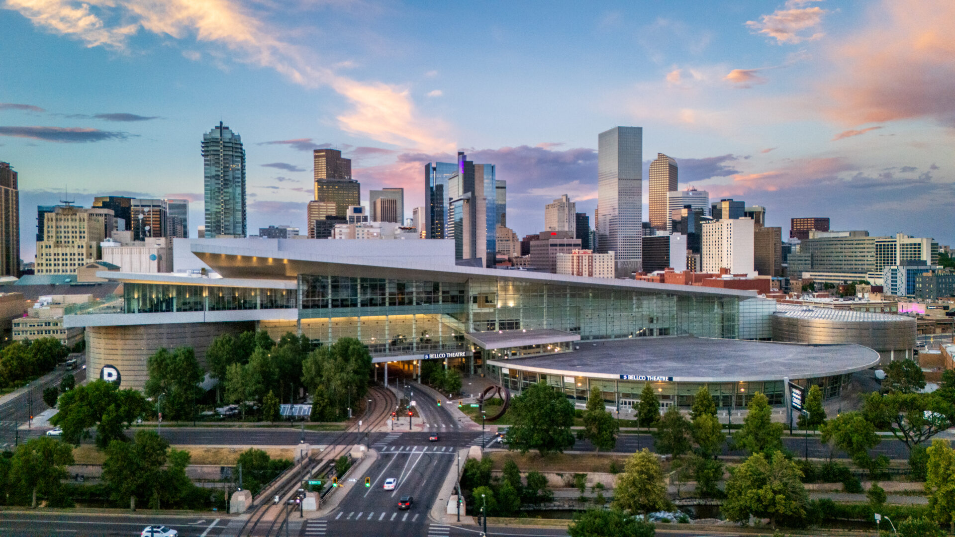 Exterior view of the Colorado Convention Center in downtown Denver, featuring its modern glass facade, the Bellco Theatre, and the city skyline beneath a colorful sunset sky.