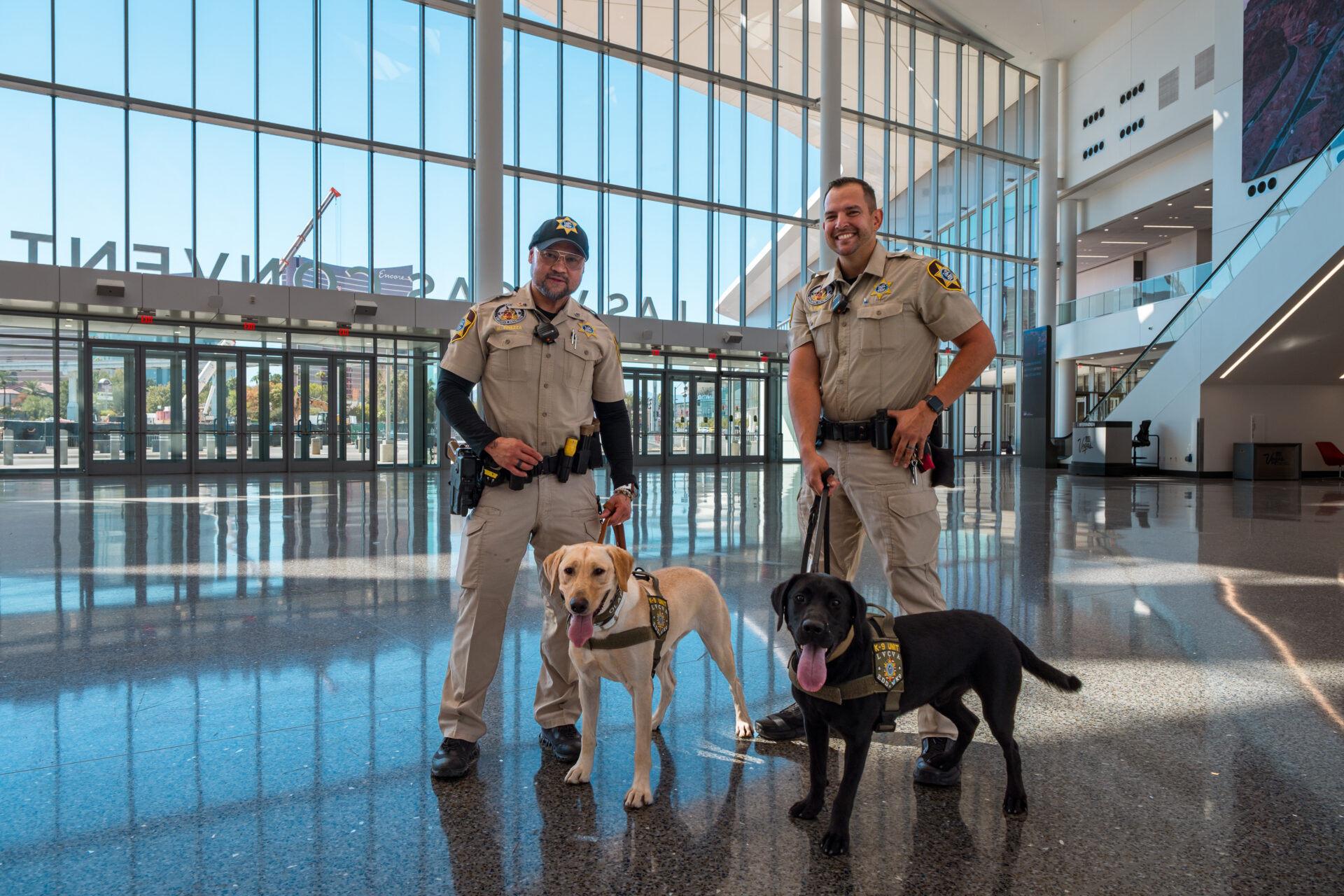Two K-9 officers stand with Olaf, a yellow Labrador Retriever, and Mufasa, a black Labrador Retriever, inside the Las Vegas Convention Center.
