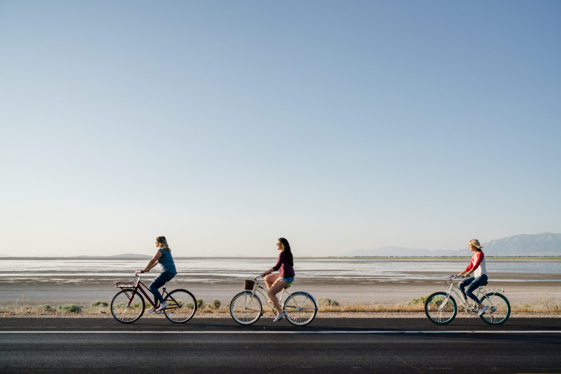 Three women ride bicycles along a paved road beside the Great Salt Lake, with expansive salt flats, distant mountains, and a clear blue sky in the background.