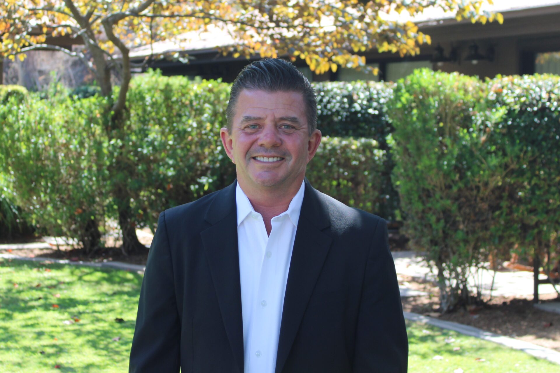 RJ Bear stands outdoors in a landscaped garden, smiling in a black suit jacket and white dress shirt.